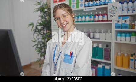 Eine lächelnde junge Apothekerin im weißen Mantel steht in einer gut organisierten Drogerie mit Regalen. Stockfoto