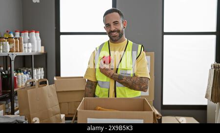 Gutaussehender hispanischer Mann mit Bart, der sich freiwillig in einem Indoor-Lagerhaus meldet, ein rotes Herz hält und Positivität projiziert. Stockfoto