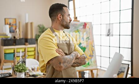 Ein selbstbewusster hispanischer Mann mit Bart trägt eine Schürze in einem hellen Kunststudio und blickt nachdenklich weg. Stockfoto