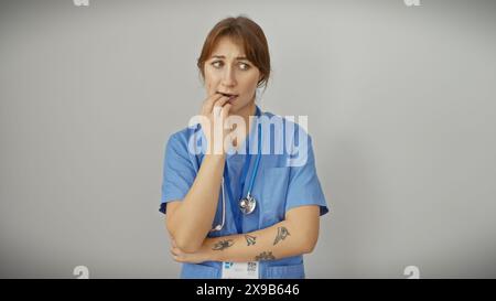 Eine besinnliche junge Frau in blauen medizinischen Peelings mit Tattoos und einem Stethoskop steht vor einem weißen Hintergrund. Stockfoto