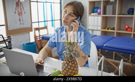Eine junge Frau in einem blauen Hemd spricht am Telefon, während sie an ihrem Laptop in einem physiotherapeutischen Klinikzimmer arbeitet. Stockfoto
