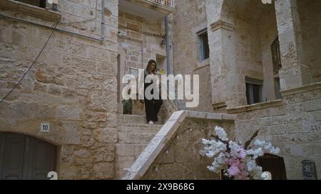 Die junge hispanische Frau in stilvollem Outfit geht die Steintreppen hinunter in den bezaubernden Straßen von polignano A Mare, apulien, italien, umgeben von einem historischen Bogen Stockfoto