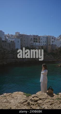 Eine junge hispanische Frau steht auf einer felsigen Klippe mit Blick auf das Meer in polignano a Mare, apulien, italien, mit malerischen Gebäuden im Hintergrund. Stockfoto