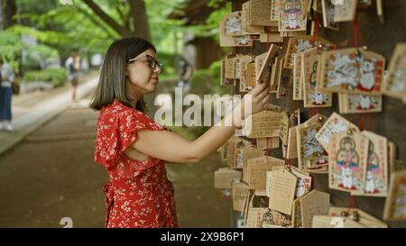 Das wunderschöne hispanische Mädchen trägt eine Brille und drückt seine Wünsche auf traditionellen japanischen Holzbrettern im Gotokuji-Tempel aus, dem Wahrzeichen der Glückskatzen Stockfoto