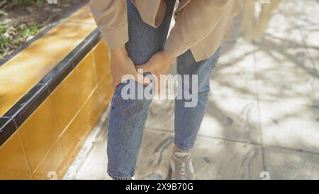 Eine junge Frau hält ihr Knie im Freien in lässiger Jeans und einer beigefarbenen Jacke, was auf mögliche Beschwerden oder Schmerzen in einer sonnigen städtischen Umgebung hinweist. Stockfoto