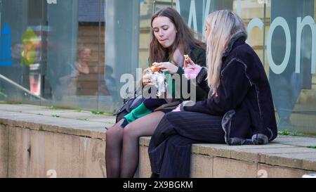 Cardiff, Wales, 29. Oktober 2023: Zwei attraktive Mädchen essen ihr Lunchpaket auf einer Steinstufe im Stadtzentrum von Cardiff. Sie unterhalten sich intensiv Stockfoto