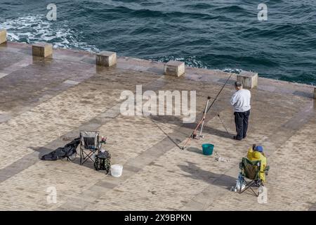 Zwei Frauen gehen, eine sitzend und ein Mann legt die Angelrute im Hafen an der Promenade des Weihnachtsleuchtturms in Cartagena. Stockfoto