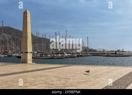 Foto einer Frau, die entlang der Esplanade des Hafens der Stadt Cartagena zwischen den beiden Säulen läuft, mit Booten, die im Hintergrund verankert sind, Murcia. Stockfoto