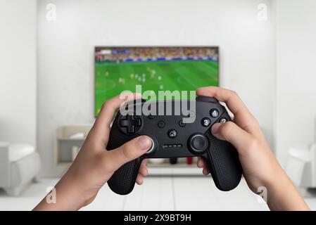 Hände halten ein Gamepad, spielen ein Fußballspiel auf einem großen Fernsehbildschirm. Perfekt für Spiele, Sport und Entertainment Stockfoto