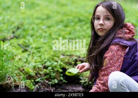 Ein kleines, wunderschönes, lächelndes, langhaariges dunkelhaariges Mädchen in Brille und Weste sitzt in der Nähe eines Baches im Wald Stockfoto