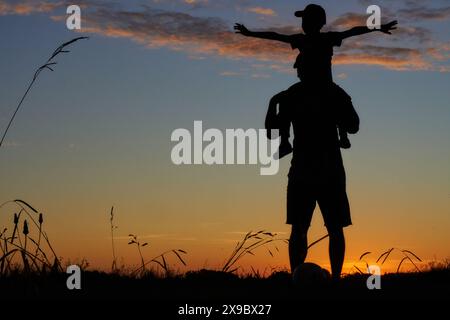 Die Silhouetten des Vaters tragen seinen Sohn auf dem Rücken, der bei Sonnenuntergang steht. Das Konzept des Vatertags Stockfoto