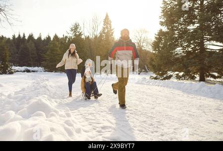Eltern nehmen Kinder mit auf Schlitten, während sie fröhlich auf verschneiten Pfaden im Winterpark spazieren. Stockfoto