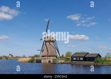 Ländliche Szene mit einer alten Windmühle auf einem See und einem kleinen blauen Haus unter klarem Himmel, Windmühlen von Kinderdijk an einem Fluss, Kinderdijk, Niederlande Stockfoto
