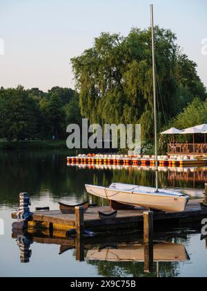 Ein Segelboot liegt an einem Steg auf einem ruhigen See, umgeben von üppigen Bäumen und reflektierendem Wasser während der Dämmerung, Steg an einem kleinen See mit Segelbooten und Stockfoto