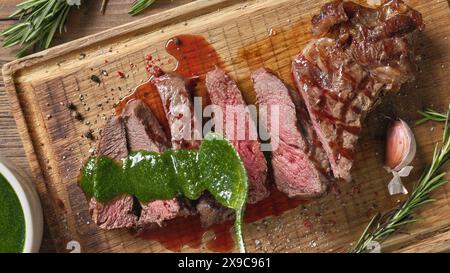 Der Koch gießt grüne Petersiliensauce auf einem hölzernen Schneidebrett auf ein frisch gegrilltes Rindfleisch. Nahaufnahme des Essens, Blick von oben Stockfoto