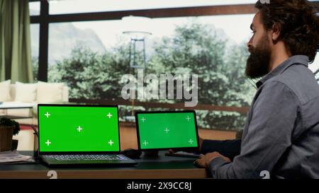 Wohlhabende Person, die auf zwei Geräten Greenscreen verwendet und an seinem Arbeitsplatz in seinem modernen, eleganten Ferienhaus sitzt. Inhaber und Investor eines reichen Unternehmens, der sich Bildschirme mit leerem Chromakey ansieht. Stockfoto