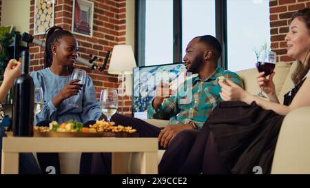 Interrassisches Paar bei einem Treffen, das auf der Couch spricht, während es ein Glas Wein genießt. Ehemann und Ehefrau Gäste auf der Apartmentfeier, die sich in einem Haus mit Ziegelmauern unterhalten und Snacks essen Stockfoto