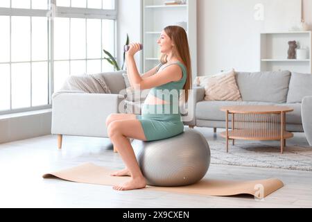 Sportliche junge schwangere Frau, die zu Hause mit Kurzhanteln am Fitball trainiert Stockfoto