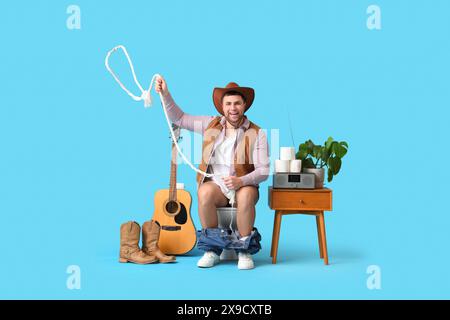 Junger Cowboy mit Seil, der auf einer Toilettenschüssel vor blauem Hintergrund sitzt Stockfoto