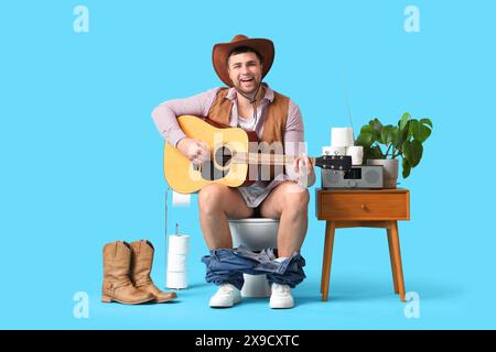 Junger Cowboy spielt Gitarre auf der Toilettenschüssel vor blauem Hintergrund Stockfoto