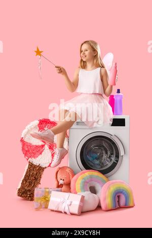 Kleines Mädchen im Feenkostüm mit Zauberstab und Flasche Waschmittel, das auf der Waschmaschine neben Geschenken vor rosa Hintergrund sitzt Stockfoto