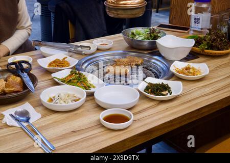 Ulsan, Südkorea - 24. Februar 2024: Grillfleisch auf einem Holzkohlegrill, umgeben von Beilagen aus Banchan, darunter Kimchi, Sojasprossen, Zwiebeln i Stockfoto