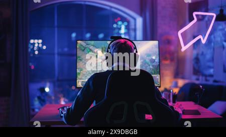 Hinterkopf-Winkel einer jungen weiblichen Gamerin, die Kopfhörer trägt und ein Videospiel auf einem PC in einem neonbeleuchteten Wohnzimmer zu Hause spielt. Gemütlicher Abend zu Hause im Loft Apartment. Stockfoto