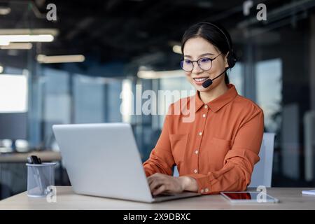 Asiatische Frau mit Headset-Telefon lächelnd im Büro mit Laptop, Online-Kundendienstmitarbeiter, Geschäftsfrau, die auf der Tastatur schreibt, Kunden aus der Ferne hilft, erfolgreiche Frau. Stockfoto