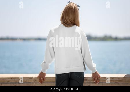 Weiße Sweatshirt-Vorlage mit seitlichen Schlitzen auf Mädchen, Rückansicht, Dame auf dem Kai, verschwommener Flusshintergrund. Leeres modisches Modell mit langen Ärmeln f Stockfoto