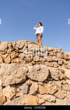 Eine Frau mit 30 in Sonnenbrille, die in lässigem Outfit nach vorne blickt, steht selbstbewusst auf alten Steinmauern vor einem klaren blauen Himmel. Lifestyle Co Stockfoto