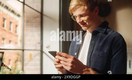 Sonniger Tag: Filmisches Foto des glücklichen schönen weißen Mannes mit Smartphone im gemütlichen Wohnzimmer zu Hause. Online-Shopping, Internet-Browsing und Social Media. Stockfoto