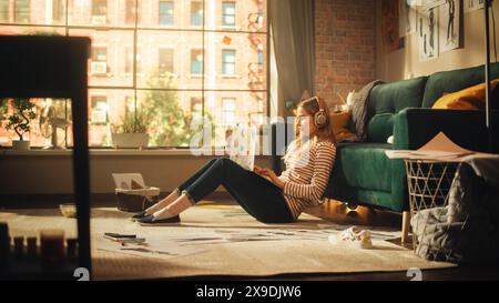 Junge hübsche Frau mit blondem Haar, die Kopfhörer trägt. Arbeiten von zu Hause aus mit einem Laptop in einem stilvollen Loft Apartment. Kreative Frau, die in entspannter Pose sitzt und soziale Medien überprüft. Stockfoto