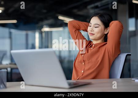 Asiatische Frau, gut gemacht, erfolgreich abgeschlossen das Projekt, Geschäftsfrau mit Händen hinter Kopf, ruht am Arbeitsplatz, Frau mit geschlossenen Augen träumend und dösen, Firmenangestellte im Büro. Stockfoto