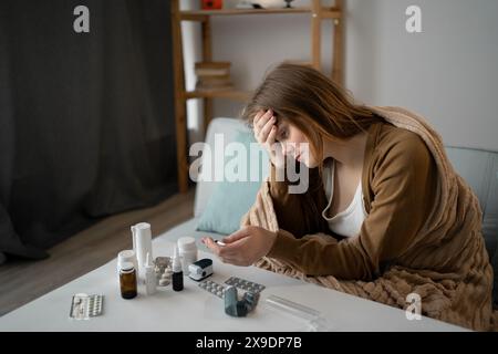 Eine kranke Frau ist auf dem Sofa in eine Decke gewickelt. Leiden an grippeähnlichen Symptomen, einschließlich Kopfschmerzen und laufende Nase. Schaut auf das Thermometer und Stockfoto