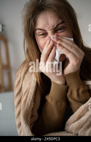 In eine Decke gehüllt, sitzt eine Frau zu Hause auf der Couch und kämpft gegen eine saisonale Erkältung. Mit einem Taschentuch in der Hand bläst sie sich zart in die Nase Stockfoto