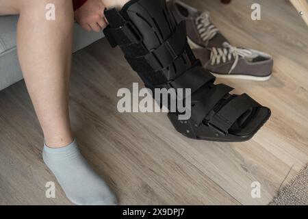 Verletzte Frau mit schwarzer Schiene am Bein sitzt zu Hause auf dem Sofa. Nahaufnahme der Knöchelstütze Stockfoto