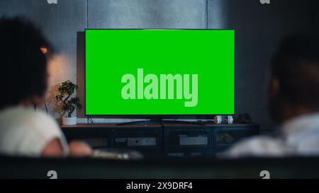 Rückansicht des glücklich verheirateten Schwarzen Paares, das Popcorn auf dem Sofa isst und Fernsehen mit Green Screen Mock Up Display im Loft Wohnzimmer sieht. Außer Fokus Paar Nahaufnahme in der Nacht. Stockfoto