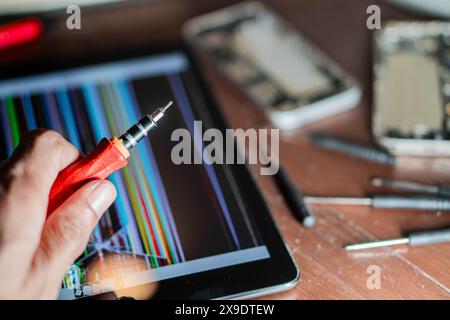 Nahaufnahme einer Person, die einen Schraubendreher in der Nähe eines Tablets hält, mit einem beschädigten Bildschirm, der bunte Linien zeigt. Verschiedene Werkzeuge und elektronische Geräte sind sca Stockfoto