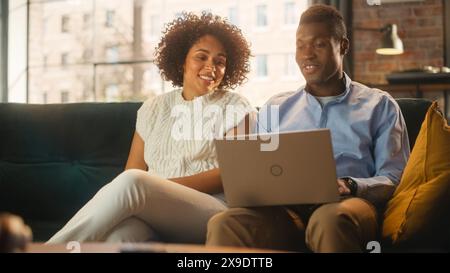 Glückliches junges Paar, das auf dem Sofa in ihrem stilvollen Loft Apartment sitzt und einen Laptop benutzt. Schwarzer Freund und Freundin diskutieren Nachrichten, während sie sich den Gerätebildschirm ansehen. Stockfoto
