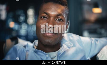 Porträt des jungen gutaussehenden Schwarzen Mannes, der über etwas lustiges im Fernsehen lächelt und lacht, während er abends auf einem Sofa sitzt. Positive menschliche Emotionen im Wohnzimmerkonzept. Stockfoto