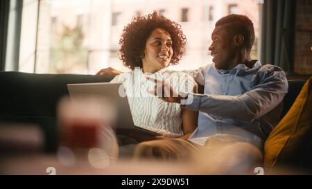 Ein multiethnisches Paar sitzt auf der Couch im stilvollen Wohnzimmer und wählt Artikel aus, die online mit einem Laptop gekauft werden können. Ein schwarzer Mann, der seine Frau mit Liebe und Unterstützung umschließt. Black Family Business. Stockfoto