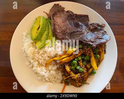 Blick von oben auf eine Platte mit gegrilltem Rindfleisch, weißem Reis, Avocadoscheiben und gebratenem Gemüse mit Erbsen, serviert auf einem weißen Teller. Stockfoto