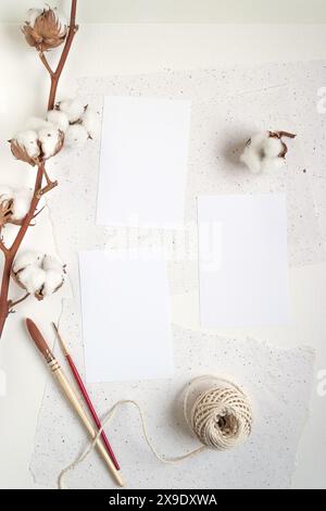 Tischmockup mit drei vertikalen leeren weißen Karten, verschiedenen Papiersorten, Pinseln und Baumwoll-Pflanzenzweigen Stockfoto