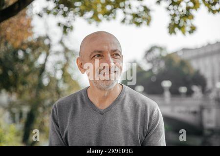 Ein älterer Mann lächelt warm, während er in einem Park, in Bäumen und im Fluss sitzt Stockfoto