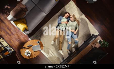 Top View Apartment: Verliebtes Paar, das sich auf einem Teppich im Wohnzimmer entspannt, mit Smartphone und Blick auf die Kamera. Freundin und Freund machen Online-Shopping, E-Business zusammen. Stockfoto
