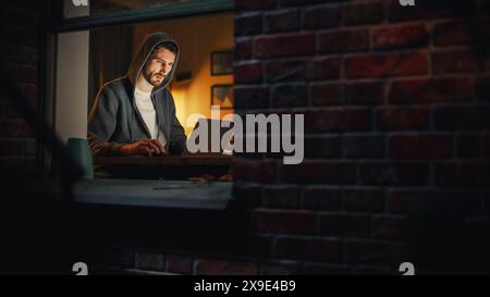 Informatikspezialist arbeitet spät an einem Latptop. Software Engineer trägt Hoodie und arbeitet am Computer in seinem gemütlichen Home Office Apartment. Fernarbeit. Nachtsicht von draußen ins Fenster. Stockfoto