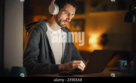 IT-Spezialist arbeitet spät an Latptop. Software Engineer trägt Hoodie und tippt am Computer in seinem gemütlichen Home Office Apartment. Fernarbeitskonzept. Blick Von Draußen Ins Fenster. Stockfoto