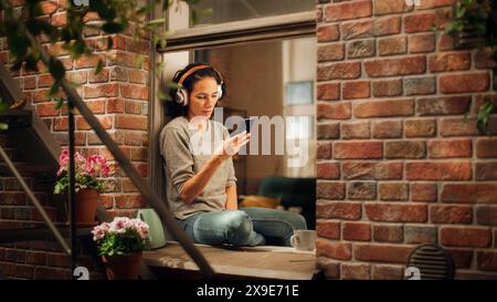 Schöne lächelnde brasilianische Frau, die auf der Fensterbank sitzt und Smartphone-Apps nutzt, Internet surft, Informationen sucht, Dating-Service, lächelt glücklich. Durch Das Fenster Des Sunny Apartments Geschossen. Stockfoto