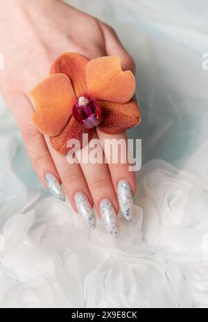 Eine Nahaufnahme zeigt eine Person Hand mit komplizierten, floralen Nailarten, die zart eine Orangenblüte umhüllt. Die umliegende weiche, schwungvolle Textur Stockfoto