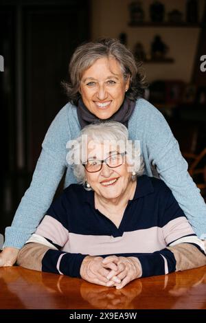 Porträt einer leichtsinnigen Frau, die an einem Tisch sitzt und warm lächelt, während ihre Tochter mit liebevoller Umarmung hinter ihr steht, die Freude und ein ausdrückt Stockfoto